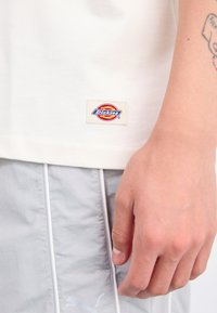 T-shirt bianca in cotone con etichetta Dickies cucita all'orlo, abbinata a pantaloni corti grigi chiari con piping bianco e logo.