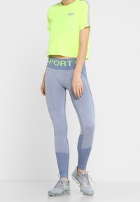 Neon żółty skrócony t-shirt z nadrukowanym logo na klatce piersiowej, w połączeniu z szarymi legginsami z niebieskimi akcentami i grubym pasem z napisem "SPORT".