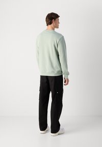 Uomo in piedi rivolto di spalle, indossa un felpa verde chiaro, pantaloni cargo neri e sneakers bianche su uno sfondo bianco uniforme.