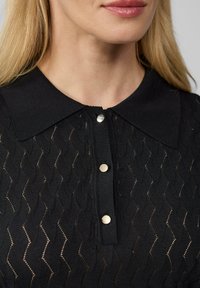 Zwarte gebreide polo met een geweven zigzagpatroon, voorzien van een kraag en drie metalen knoopaccenten aan de voorkant.