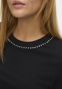 Maglietta nera con scollatura rotonda decorata da una fila di piccoli borchie argentate lucide attorno al colletto. Texture liscia.