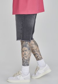 Pantalones cortos de mezclilla negra con dobladillos deshilachados, combinados con zapatillas blancas y calcetines blancos. Tatuajes visibles con diseños intrincados y retratos.