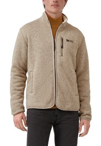 Mann trägt eine beigen Fleecejacke mit Reißverschluss über einem senfgelben Rollkragenpullover, mit einer Reißverschlusstasche auf der Brust und entspannten Händen an den Seiten.