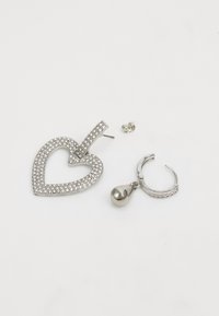 Zilveren oorbelhanger in de vorm van een hart met rhinestones, naast een zilveren ringoorbel met een druppelaccent. Gemaakt van metaal.
