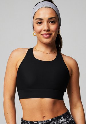 Frau trägt einen schwarzen Sport-BH, gemusterte Trainingsshorts, ein graues Stirnband und goldene Creolen, lächelnd vor einem einfarbigen Hintergrund.