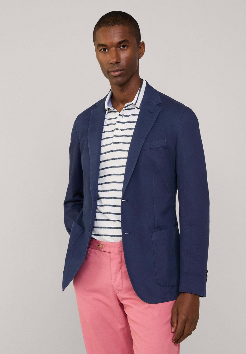 Granatowa marynarka z szpiczastymi klapami i dwoma przednimi kieszeniami, zestawiona z paskowanym polo i jasnoróżowymi spodniami. Dopasowany krój, materiał bawełniany.