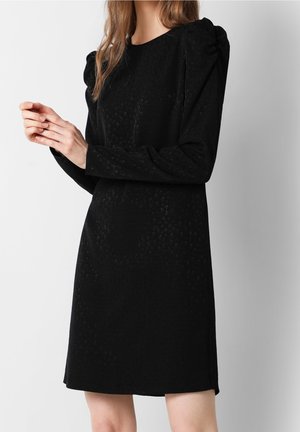 Femme portant une robe noire à manches longues avec un motif texturé subtil et des épaules légèrement bouffantes, debout devant un fond clair uni.