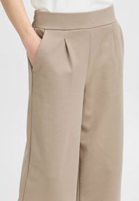 Pantalons large beiges avec des plis, fabriqués en tissu léger. Présente une taille haute avec des coutures verticales et des poches latérales.