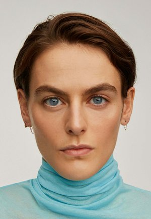 Persona con cabello corto castaño y ojos azules, vistiendo un suéter de cuello alto color azul claro y pequeños pendientes de aro dorados, con expresión facial neutral.