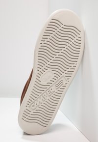 Zapato marrón con una suela de goma blanca flexible que presenta patrones de tracción ondulados y la marca "Camper". El zapato está hecho de material suave y tiene una forma redondeada.