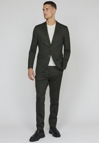 Dark green suit with a tailored blazer and matching trousers, featuring a smooth texture and single chest pocket. Paired with a white t-shirt.