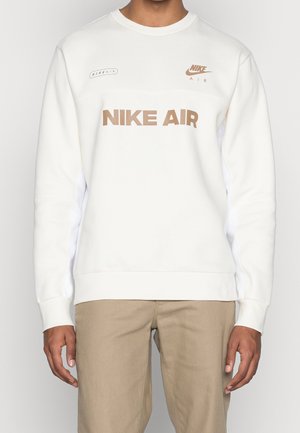 Personne portant un sweat-shirt blanc Nike Air avec un logo beige et un pantalon beige, debout les bras détendus le long du corps.