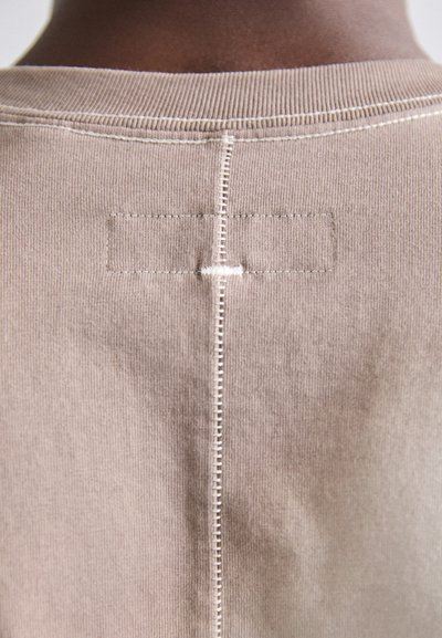 Vêtement en coton marron clair avec une couture verticale dans le dos, avec des coutures visibles et un petit patch rectangulaire au-dessus de la couture.