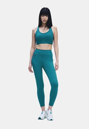 Junge Frau steht in einem türkisfarbenen Sport-BH und passenden hoch taillierten Leggings mit weißen Sportschuhen vor einem schlichten Hintergrund.