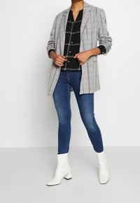 Grau karierter Blazer mit Revers über einem schwarzen Button-Up-Hemd, kombiniert mit blauen Skinny-Jeans und weißen Blockabsatz-Stiefeletten.