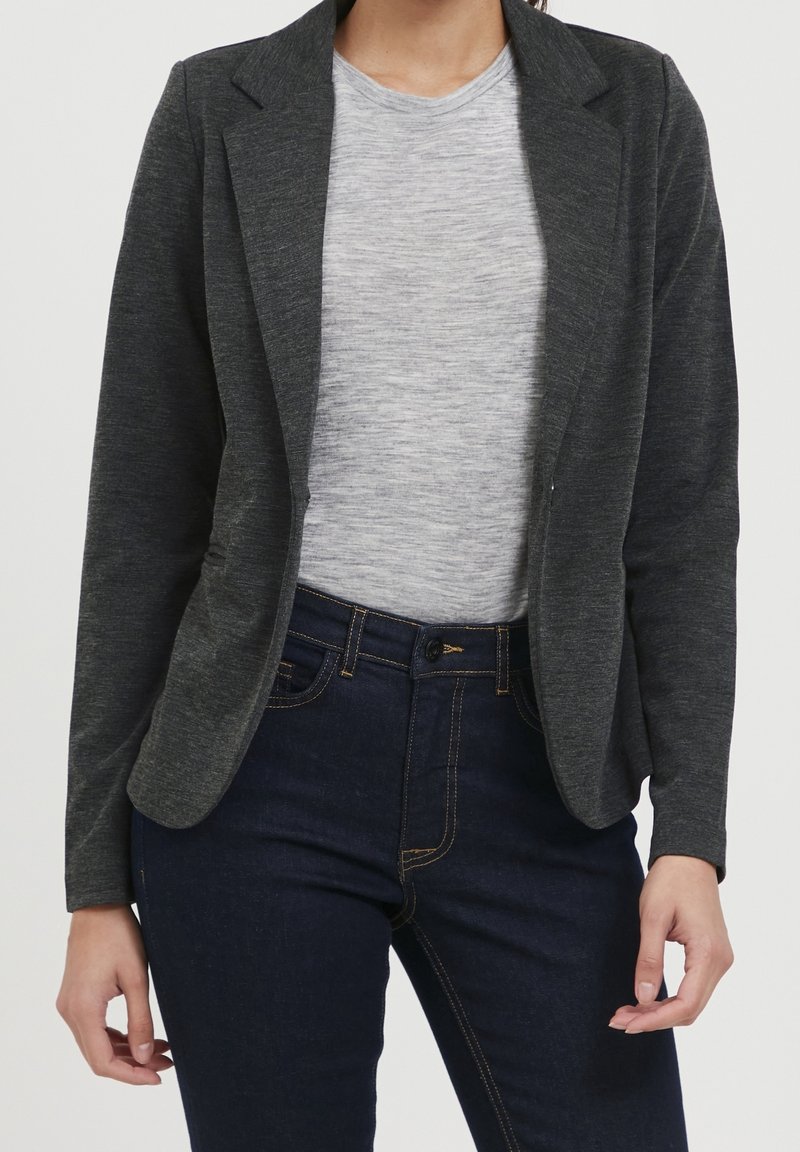 Tmavě šedý blazer s přiléhavým střihem, zapínáním na jeden knoflík a klopovými límci, v kombinaci se světle šedým tričkem a tmavě modrými džíny.