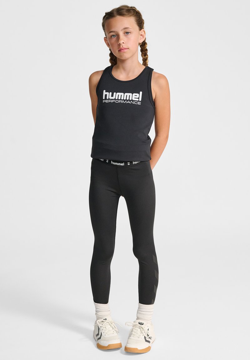 Schwarzes ärmelloses Oberteil mit "hummel PERFORMANCE"-Logo; schwarze Leggings mit seitlichen Details; weiße Sneaker mit beigen Akzenten; helle Socken.
