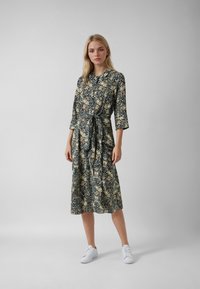 Vestito a fiori blu scuro con dettagli beige, caratterizzato da una vita regolabile, maniche a tre quarti e lunghezza midi, abbinato a scarpe da ginnastica bianche.