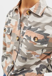 Camicia camouflage con motivi beige, grigi e corallo, due tasche frontali, bottoni marroni tondi e colletto a punta in un tessuto strutturato.