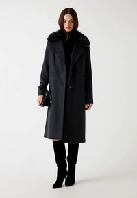 Długi czarny wełniany płaszcz z szerokim kołnierzem z futerka ekologicznego, dwoma przednimi kieszeniami i prostym krojem. Noszony z czarnym golfem i wysokimi do kolan kozakami.