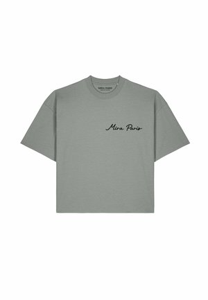 Frau trägt Sonnenbrille und graues Shirt mit abstraktem Gesichtsmotiv, steht mit der Hand in der Tasche in der Nähe des Eiffelturms in Paris.