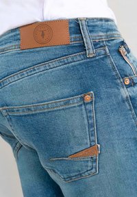 Jeans en denim bleu avec un patch en cuir marron à l'arrière, des coutures oranges, une fermeture éclair et un détail de poche sur le côté droit.