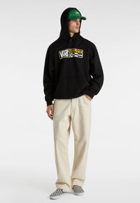 Zwarte hoodie met Vans-logo in geel en wit, crème kleurige broek en zwart-witte geruite slip-on schoenen. Groene pet maakt de look compleet.