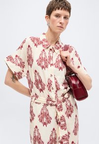 Femme aux cheveux courts portant une robe beige avec des motifs cachemire rouges, tenant un sac à main en cuir rouge sur l'épaule, debout devant un fond uni.