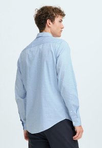 Cămașă bleu deschis, cu un model geometric mic, mâneci lungi și o tivire curbată. Fabricată dintr-un material texturat, cu nasturi albi.