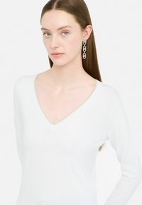 Pulover alb cu decolteu în V, cu o margine din fir argintiu, realizat dintr-un material moale. Modelul poartă cercei lungi din lanț argintiu. Design simplu și elegant.