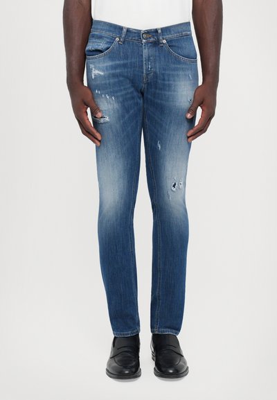 Jeans in denim blu scuro con vestibilità slim, caratterizzati da un lavaggio sbiadito, dettagli strappati e design a cinque tasche. Indossati con scarpe nere.