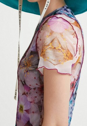 Haut court à manches courtes en tissu transparent orné de fleurs roses et orange porté par une personne avec un chapeau bleu et une sangle blanche autour du cou portant l'inscription « LIFE IS A WAVE ».