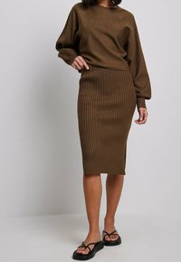 Maglione di maglia marrone con spalle abbassate, abbinato a una gonna midi marrone a coste. Il modello indossa sandali neri con plateau. Sfondo semplice.