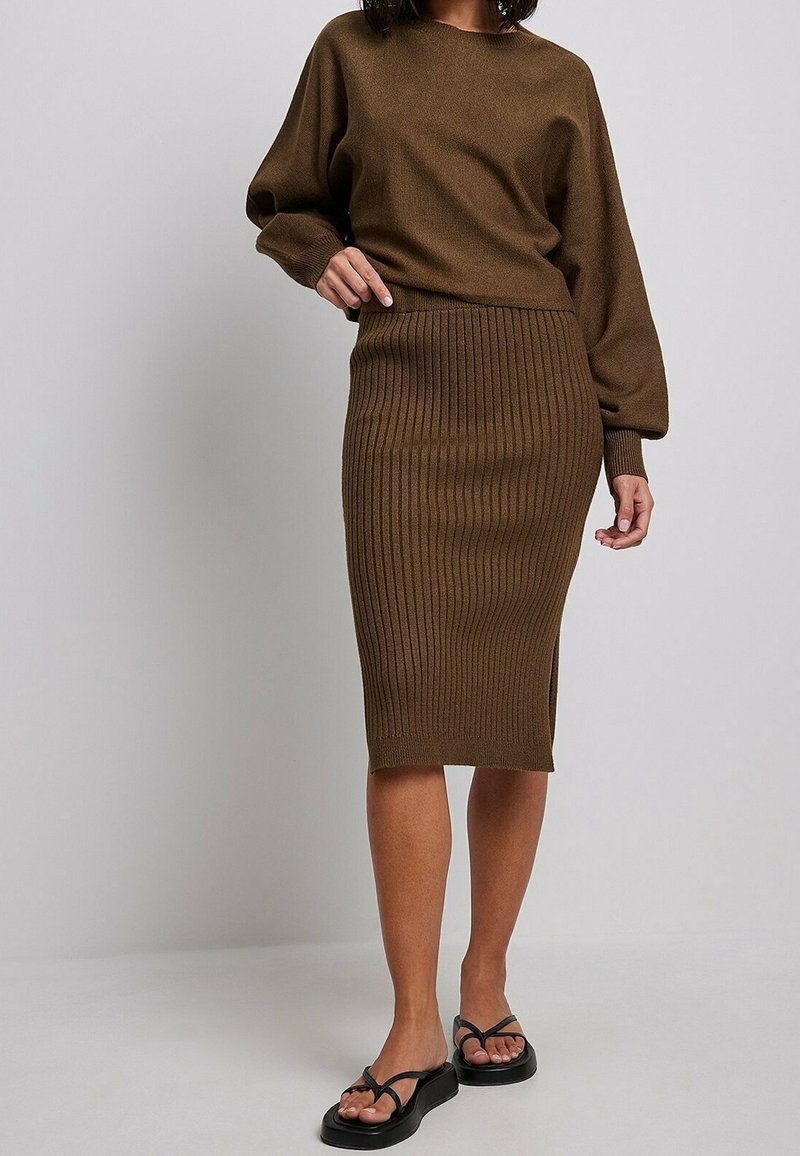 Maglione di maglia marrone con spalle abbassate, abbinato a una gonna midi marrone a coste. Il modello indossa sandali neri con plateau. Sfondo semplice.