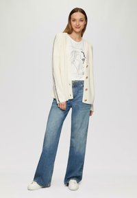 Ecru geribde cardigan met gouden knopen, gedragen over een wit graphic t-shirt. Gecombineerd met lichtblauwe wijde spijkerbroek en witte sneakers.