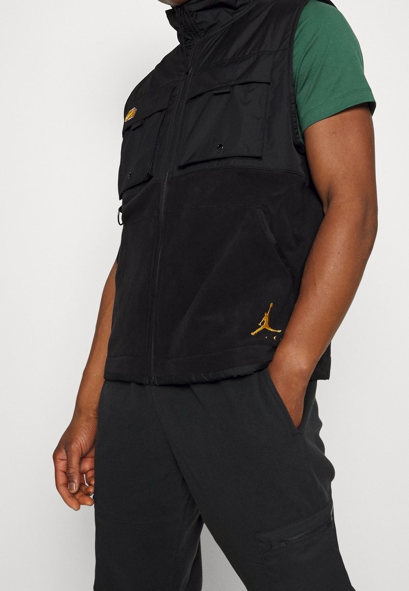 Černá vesta bez rukávů s vysokým límcem a dvěma předními kapsami. Má žlutý logo akcent a jemnou texturu. Nositelná přes zelenou košili.