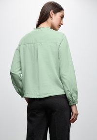 Blusa verde menta a maniche lunghe con collo arricciato, tessuto leggero e forma ampia. Sul retro presenta una piega per un maggiore movimento.