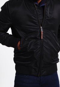 Schwarze Bomberjacke aus glattem synthetischem Material mit gerippten Bündchen und Saum, Reißverschluss mit brazem Leder-Anhänger und seitlichen Taschen.