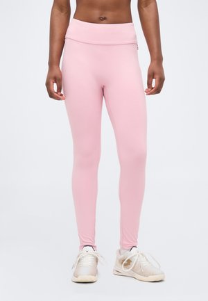 Persona en forma que lleva leggins rosa pastel de tiro alto y zapatillas deportivas blancas, de pie con las manos relajadas a los lados.