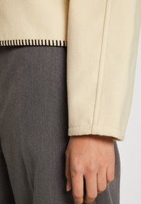 Beżowa polarowa kurtka z czystym dekoltem, widocznym czarnym przeszyciem i prostymi rękawami. Szary materiał widoczny pod spodem.