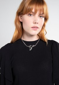 Sølv kædedesign med en abstrakt løb, præget af en poleret finish og vekslende links, båret over en sort ribbet top.