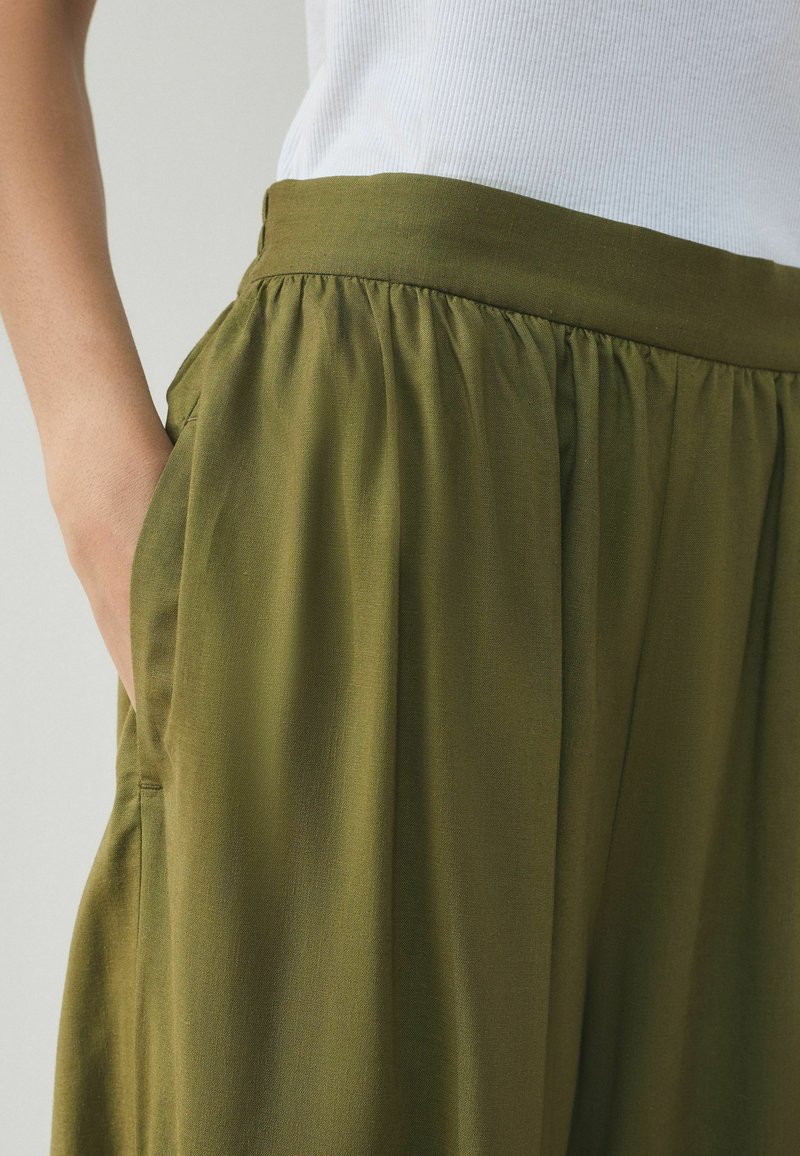 Personne portant un pantalon ample taille haute olive avec une ceinture froncée et la main dans la poche latérale, associé à un haut blanc côtelé.