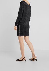 Abito nero con glitter, maniche lunghe, scollatura profonda sulla schiena e dettaglio con zip laterale. Abbinato a décolleté nere con tacco a slitta e dettagli a fiocco.