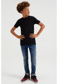 Černé tričko s krátkým rukávem spárované se sepranými modrými skinny džíny a červeno-černými teniskami. Modelka stojí s rukama na bokách.