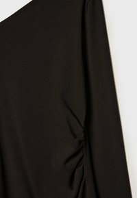 Vêtement noir à manches longues avec un détail froncé sur la couture latérale, présenté sur un fond clair.