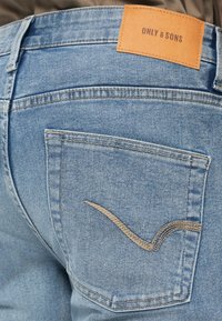 Jeans din denim de culoare albastru deschis au o buzunar din spate cu un design de cusătură curved de culoare crem și o etichetă din piele pe care scrie "ONLY & SONS."