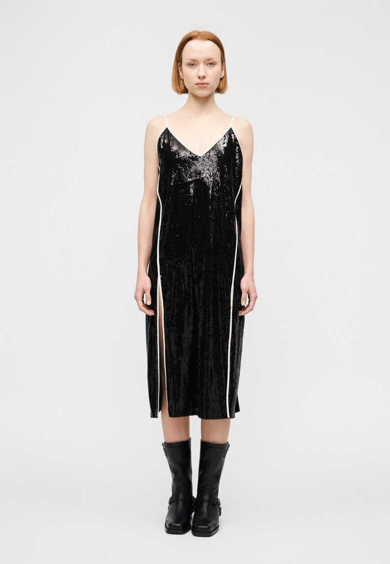 Μαύρο φόρεμα slip με παγιέτες, λευκές πλαϊνές ραφές και V-λαιμόκοψη· διαθέτει μήκος μέχρι τη μέση της γάμπας και πλαϊνές σχισμές, σε συνδυασμό με μαύρα μπότες μέχρι το γόνατο.