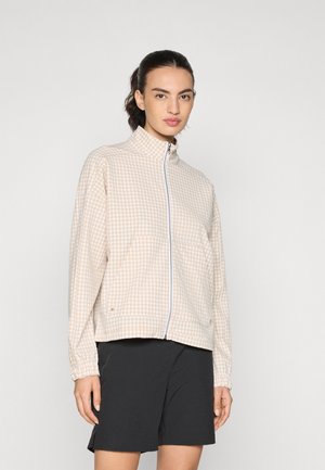 Beige ruudullinen vetoketjullinen takki, jossa on korkea kaulus, elastiset hihansuut ja kaksi etutaskua. Päällä mustat shortsit, joissa on rento istuvuus.