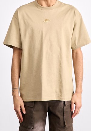 Person trägt beige Nike-T-Shirt und dunkle Cargo-Shorts, zeigt oberen Torso und Arme mit Tätowierung am linken Handgelenk.