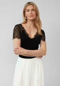 Zwart ribbel top met korte kanten mouwen en een V-hals, gecombineerd met witte hoge taille broeken. Subtiele franjes langs de kanten randen.